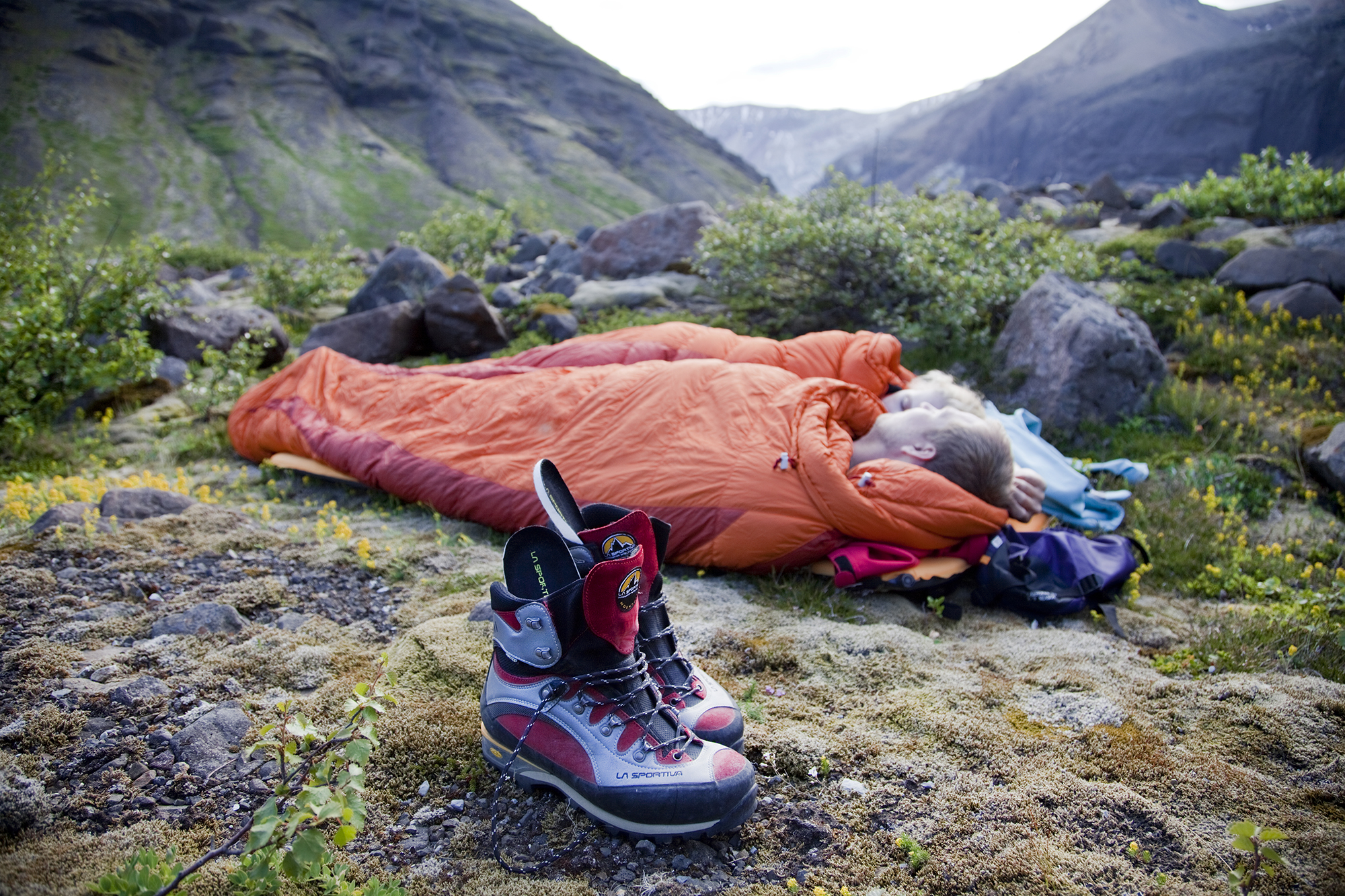 Fólk í appelsínugulum svefnpokum að lieggja sig í mosa með par af gönguskóm við hlið sér